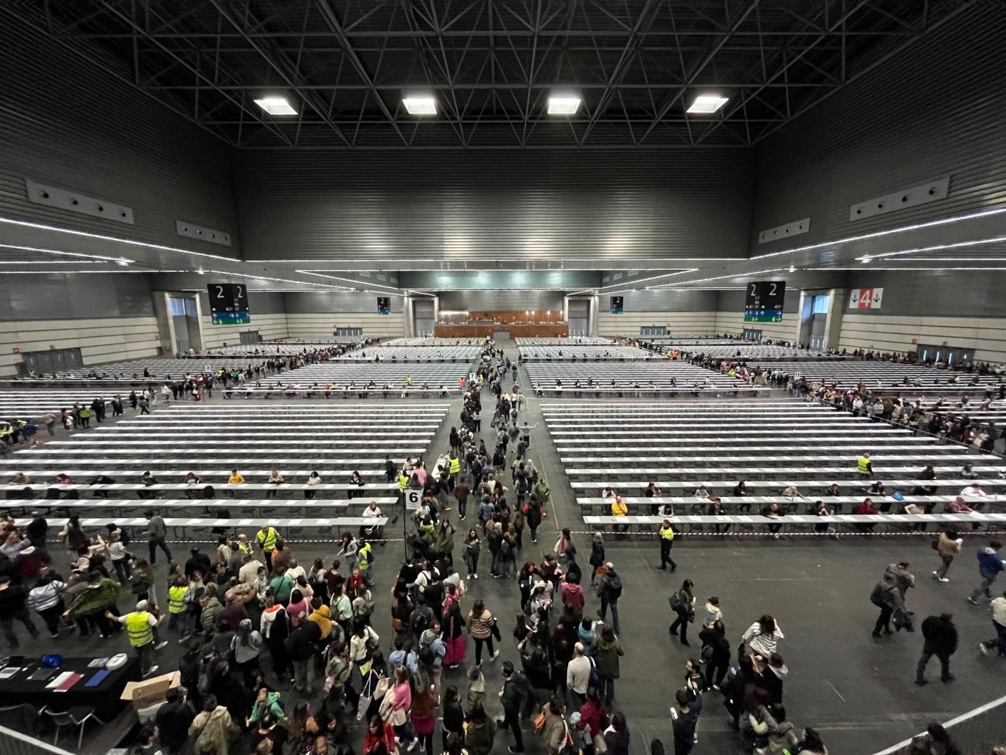 Areto erraldoi bat, mahaiak eta aulkiak lerrokatuta dituena, eta jende pila bat ilara egiten tokiren batean esertzeko. Antolatzaileek jantzi horiak daramatzate. Large convention hall with rows of long tables and many people queuing and walking between them, some wearing high-visibility vests.