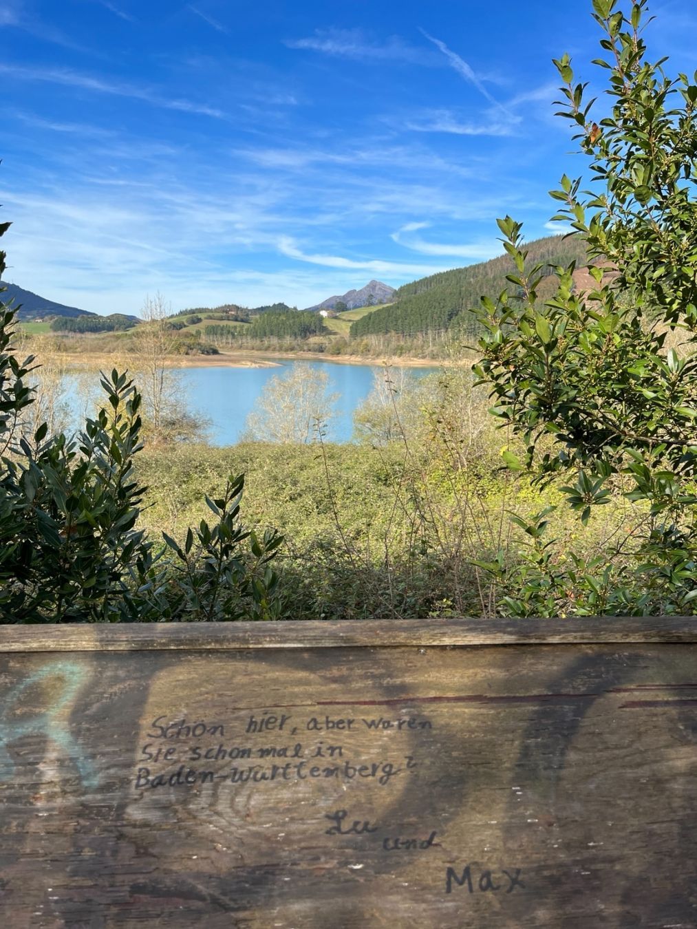 Urkulu urtegia Koruetan (Gipuzkoa). Ura zuhaitz artetik ikusten da eta zerua erabat garbi ez badago ere, urdina da egun eguzkitsuan. Argazkia ikustoki batetik eginda dago eta egurrezko taula bat dago aurrealdean. Taula horretan, errotulagailu beltzez idatzita, alemanez, Lu eta Max izeneko batzuek galdetzen digute ea, Korueta ederra izanda ere, Baden-Württembergen egon garen inoiz.