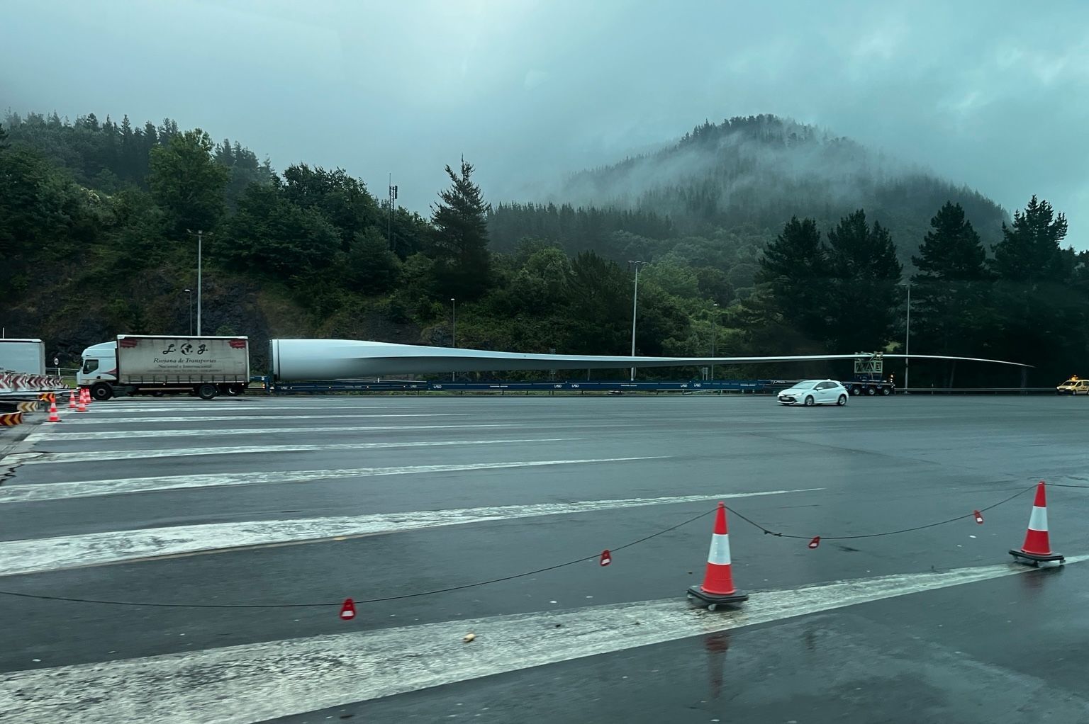 Energia sortzeko haize-errota baten paletako bat, kamioi batek daramala, AP-68 autopistako Arrigorriagako bidesarian. Giroa goibela da, errepidea hezea dago eta atzean, hondoan, baso eta mendixka bat ikusten dira laino artean.